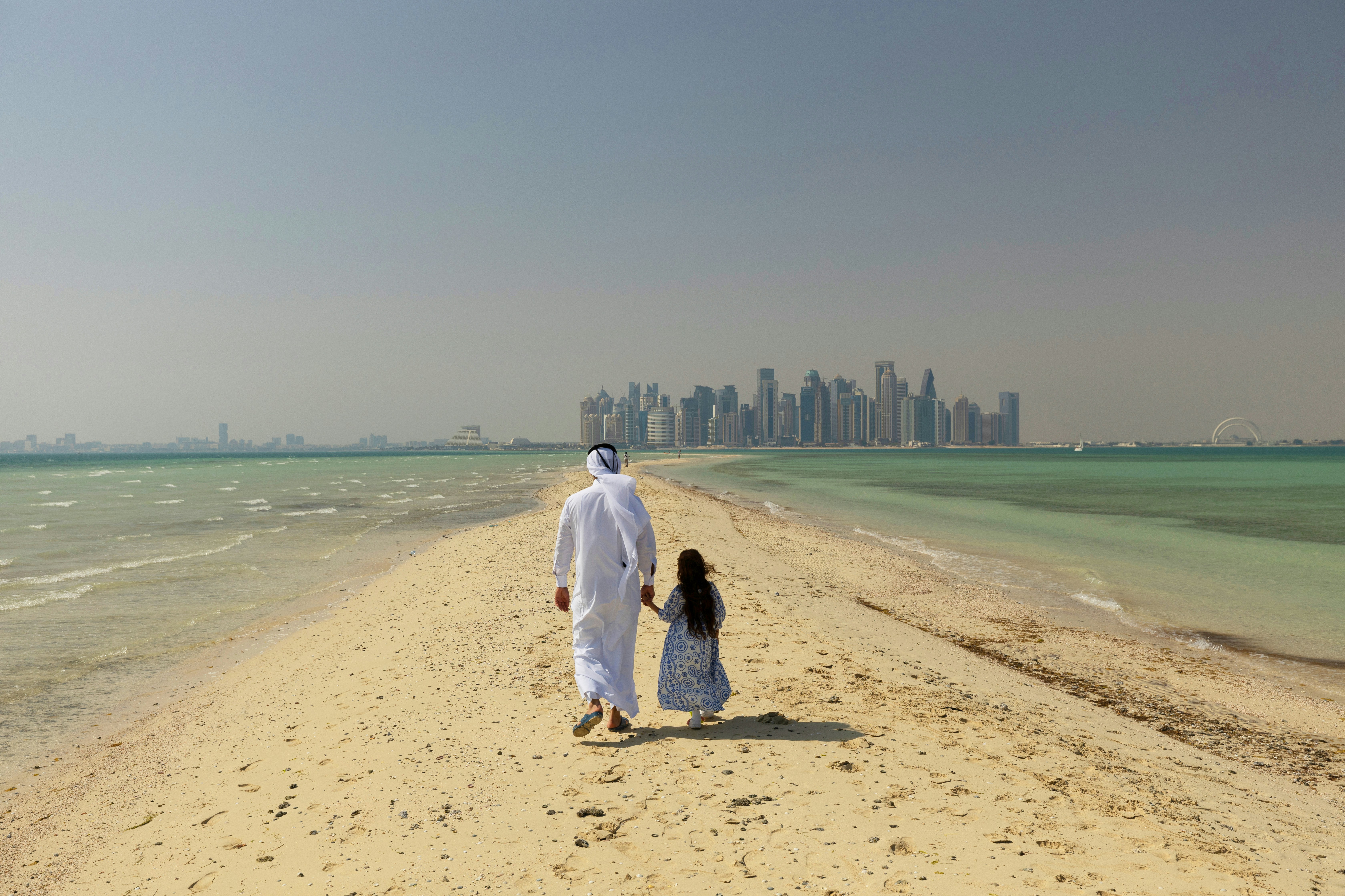 A man in traditional Gulf dress walks hand-in-hand with a young girl on a sand spit, Doha skyline in the distance
